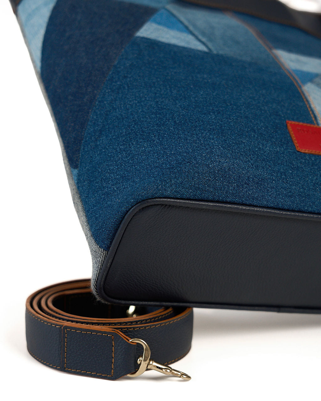 Close-up of a blue patterned fabric bag with a black leather base and a matching blue leather strap with gold-tone hardware, shown on a white background.