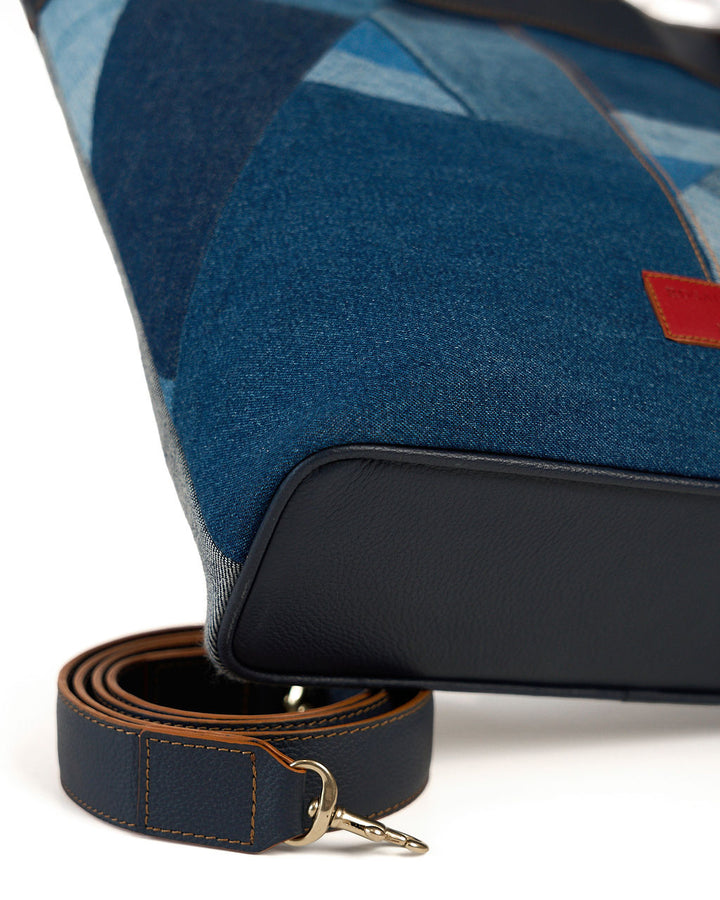 Close-up of a blue patterned fabric bag with a black leather base and a matching blue leather strap with gold-tone hardware, shown on a white background.
